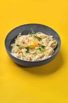 Ceramic bowl of appetizing pasta carbonara with creamy sauce, pieces of guanciale and egg yolk sprinkled with grated parmesan, presented on vibrant yellow background. Traditional Italian cuisine