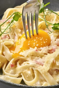 Fork gently piercing golden egg yolk on top of classic carbonara pasta, enriched with pancetta chunks and grated parmesan garnished with green pea sprouts served in gray ceramic bowl