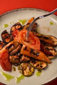Slice of juicy browned tomato on fork, on background of plate with mix of char-grilled vegetables with herb-infused oil drizzle, against red background. Healthy and delicious vegetarian dish