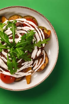 Top view of warm salad of succulent grilled vegetables and beef strips drizzled with creamy white sauce, garnished with fresh arugula on green background