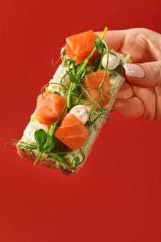 Female hand holding crispy bruschetta topped with cream cheese, pieces of cured salmon, mozzarella pearls and delicate green pea shoots against vibrant red background