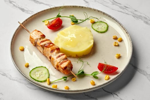 Delicious healthy food for kids menu. Mini grilled salmon skewer with portion of mashed potatoes garnished with fresh greens, corn kernels, cucumber and cherry tomato slices, against marble background