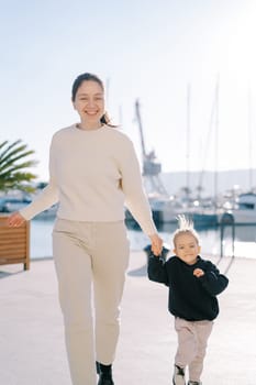 Laughing mother and little girl running along the pier holding hands. High quality photo