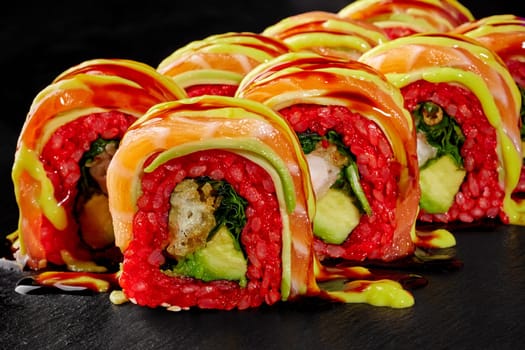 Macro shot of decadent sushi rolls with shrimp tempura, covered in slices of salmon and drizzled with savory sauces, served on black stone surface for luxurious Japanese style dining experience