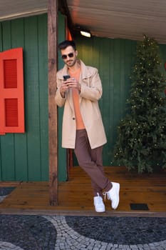Full body of stylish man wearing sunglasses and trench coat using smartphone in Christmas city decorations. Handsome young hipster standing near decorated Christmas tree in wooden house.