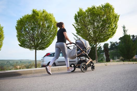 View from the back of a young woman, young mother running jogging exercising outdoors with the stroller of her baby to stay in shape after childbirth.