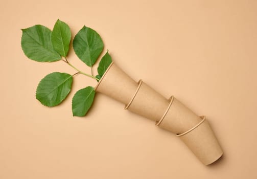 Stack of brown paper disposable cardboard cups on beige background and green leaves, top view