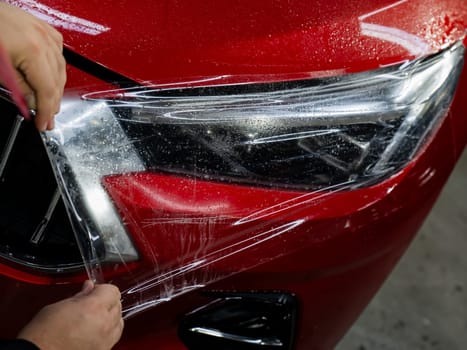 The master applies vinyl film to the headlight of a red car