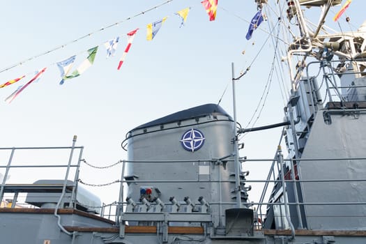 Klaipeda, Lithuania - August 11, 2023: A close-up view of a naval ships deck, showcasing a large gray gun turret with a NATO insignia, festooned with colorful signal flags under a clear sky.