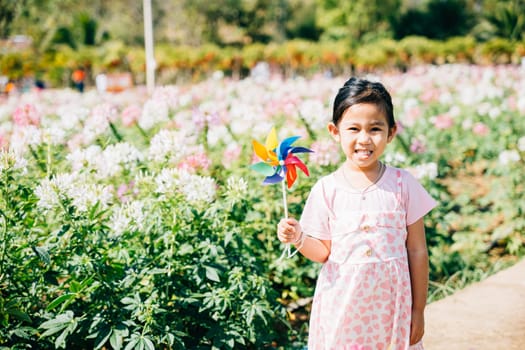 Portrait of a joyful girl in a flower garden holding a toy pinwheel. The lively spring season evokes happiness and the spinning pinwheel embodies childhood joy and freedom in nature's sunny beauty.