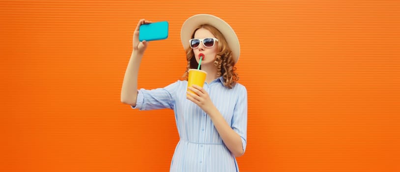 Portrait of happy relaxed young woman taking selfie with smartphone drinking fresh juice wearing a summer straw hat on orange background