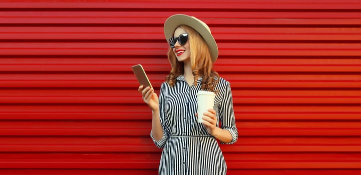 Summer portrait of stylish happy young woman with mobile phone looking at device in straw hat, holding cup of coffee on red background
