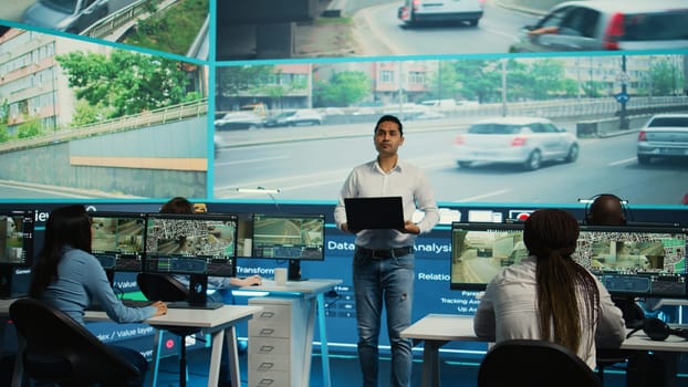 Indian team leader overseeing his employees work on surveillance footage observation, ensuring public safety via CCTV security camera around the city. Diverse government workers. Camera B.