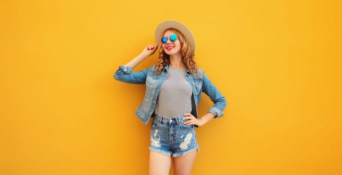 Stylish beautiful young woman wearing summer straw hat, jean jacket and shorts posing on orange background