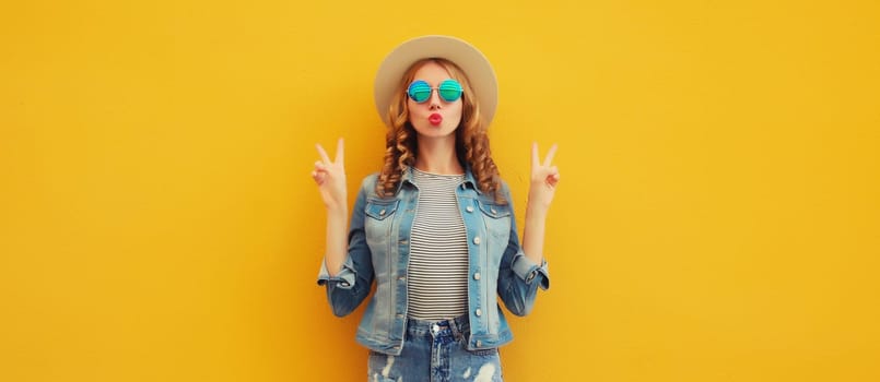 Portrait of beautiful stylish young woman blowing kiss in summer hat, sunglasses, denim jacket posing on yellow studio background