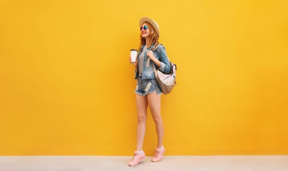 Beautiful stylish happy smiling young woman with cup of coffee wearing summer hat, backpack, jean jacket on yellow background
