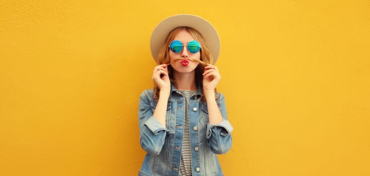 Portrait of funny young woman showing mustache her hair blowing lips with red lipstick wearing summer straw hat on orange background