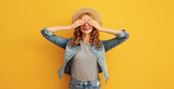 Stylish surprised young woman covering her eyes with hands wearing summer straw hat, jean jacket posing on orange background
