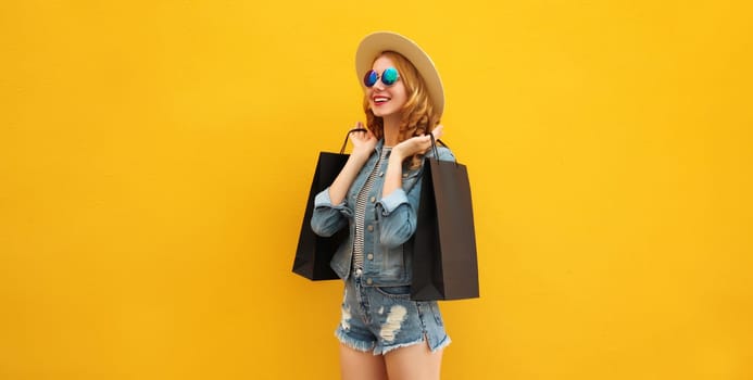 Shopping day! Stylish beautiful happy smiling young woman posing with black shopping bags wearing denim clothing, summer straw hat on yellow studio background