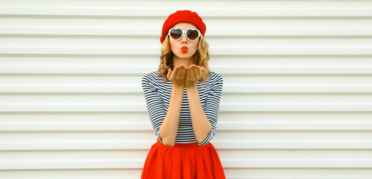 Portrait of beautiful happy young woman blowing a kiss with lipstick in red french beret, heart shaped sunglasses on white background