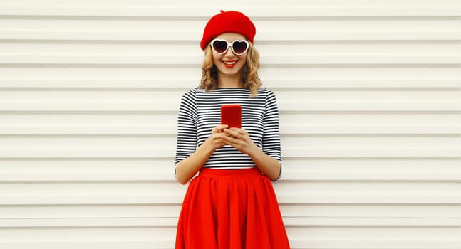 Portrait of beautiful happy smiling young woman with mobile phone looking at device wearing stylish outfit, red french beret hat, heart shaped glasses on white background