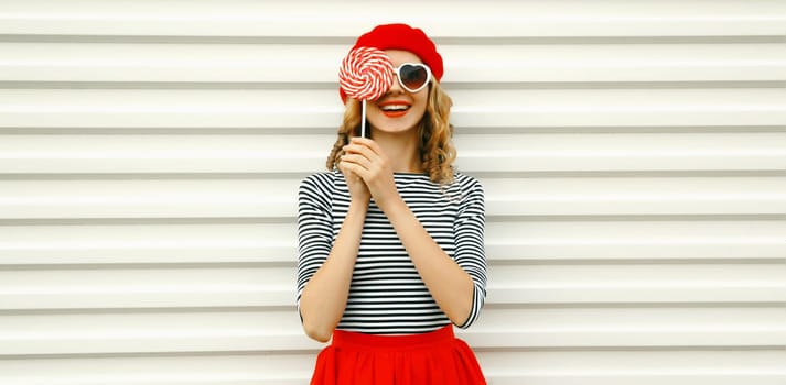 Portrait of happy cheerful smiling young woman holding colorful lollipop wearing white sunglasses, red beret hat on white background