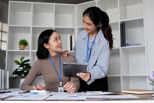 Two Asian businesswomen discussing work using a tablet in a modern office. Concept of teamwork and professional collaboration.