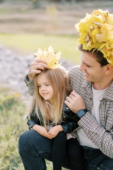 Smiling dad in a wreath of yellow leaves puts autumn leaves on the head of a little girl sitting on his lap. High quality photo