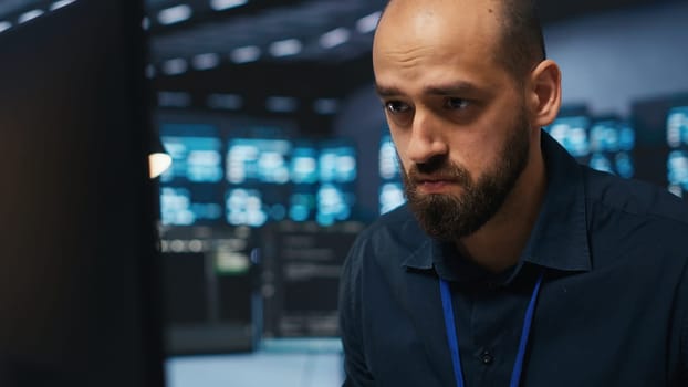 Overworked engineer feeling stressed while coding in data center facility, trying to ensure optimal performance of supercomputers. Tired man feeling fatigued while using PC to monitor server cabinets