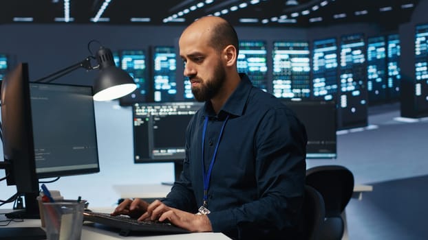Engineer coding, working in network security data center facility hosting supercomputers, ensuring optimal performance. IT expert using desktop PC to monitor energy consumption across server cabinets
