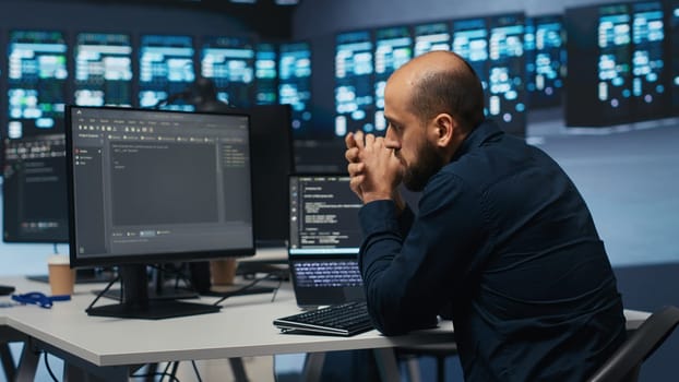 Man in server room typing code on laptop and PC, ensuring data remains shielded from threats. IT expert using notebook and computer to protect supercomputers against unauthorized access