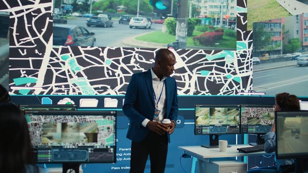 Supervisor arriving in the control center at shipping agency, overseeing main gps locating activities to ensure order and parcel delivery. Diverse employees working with satellite maps. Camera B.