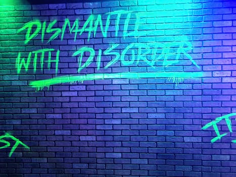 Graffiti 'Dismantle with disorder' on brick wall under neon lights in Indianapolis, echoing themes of rebellion and urban art.