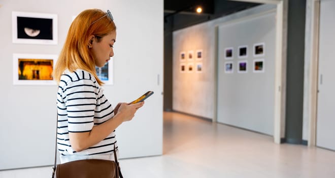 Asian woman standing takes picture art gallery collection in front framed paintings pictures on white wall with mobile phone, people watch at photo frame with smartphone at artwork gallery show