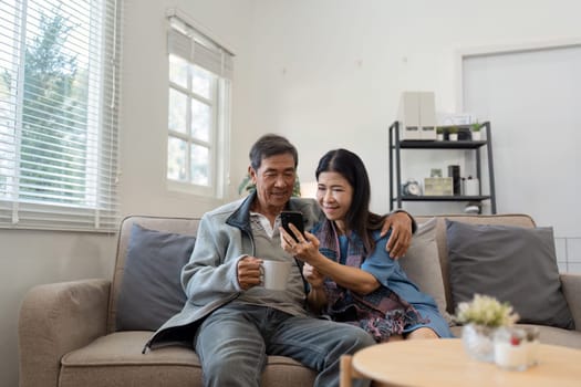 Happy married retirement couple looking at mobile using smartphone mobile technology device together, relaxing on couch doing online shopping.