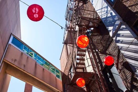 Vibrant urban alley in Fort Wayne with red lanterns and rustic pedestrian bridge against a blue sky.