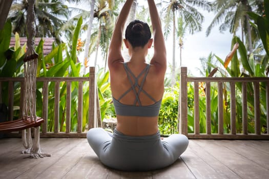 Rear view of young woman practicing yoga outdoors. Female stretching arms up in tropical vacation resort. Healthy and active lifestyle concept.