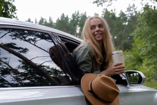 Smiling young woman looking from car window and drinking coffee or tea from reusable thermos cup. Local solo travel on weekends concept. Exited woman explore freedom outdoors in forest. Unity with nature lifestyle, rest recharge relaxation