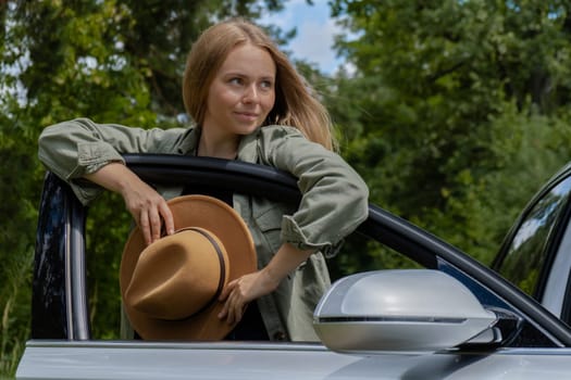 Smiling young woman in hat stoped on road to make a break in driving car. Local solo travel on weekends concept. Exited woman explore freedom outdoors in forest. Unity with nature lifestyle, rest recharge relaxation