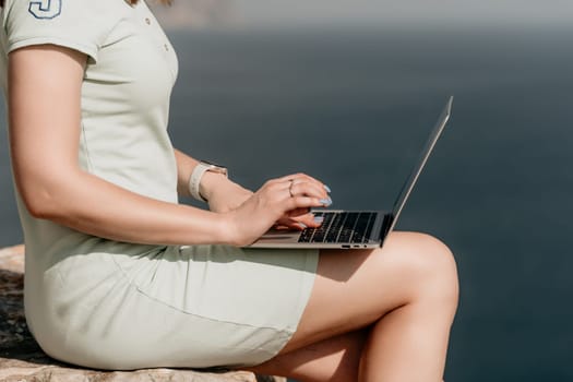 Digital nomad, woman in the hat, a business woman with a laptop sits on the rocks by the sea during sunset, makes a business transaction online from a distance. Freelance, remote work on vacation.