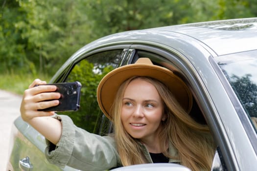 Exited smiling woman making video call with mobile phone from car window. Local solo travel on weekends concept. Young traveler explore freedom outdoors in forest taking selfie photo. Unity with nature lifestyle, rest recharge relaxation