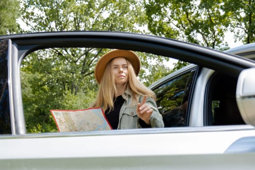 Blonde woman in hat staying next to car door checks the route on map to get to the destination. Young tourist explore local travel making candid real moments. True emotions expressions of getting away and refresh relax on open clean air