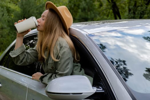 Smiling young woman looking from car window and drinking coffee or tea from reusable thermos cup. Local solo travel on weekends concept. Exited woman explore freedom outdoors in forest. Unity with nature lifestyle, rest recharge relaxation