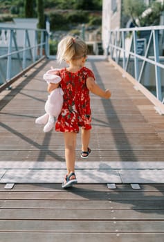 Little girl walks along the pier with a pink soft toy hare under her arm. Back view. High quality photo