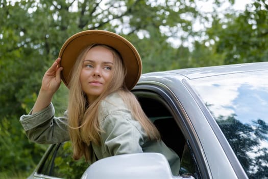 Smiling young woman in hat looking from car window. Local solo travel on weekends concept. Exited woman explore freedom outdoors in forest. Unity with nature lifestyle, rest recharge relaxation