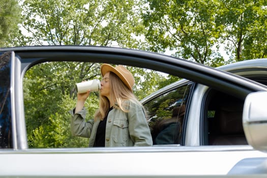 Blonde woman stoped on road next to car and drink coffee or tea from reusable mug. Refuse reuse recycle zero waste concept. Young tourist explore local travel making candid real moments. Responsible traveling reduce carbon footprint sustainable lifestyle