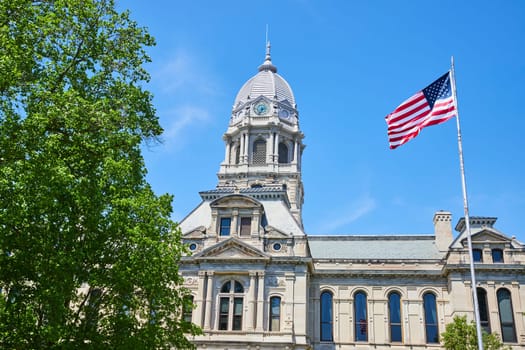 Sunny view of Kosciusko County Courthouse in Warsaw, featuring a waving American flag and lush foliage.