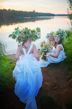 Ivan Kupala Celebration. Three Girls With Floral Wreaths by the River at Sunset. Family clad in white dresses celebrate Ivan Kupala by river at dusk.