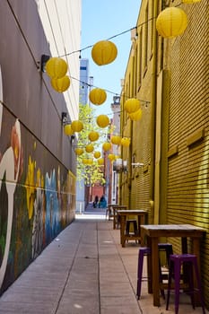 Colorful urban alley in Fort Wayne adorned with yellow lanterns and a vibrant floral mural, ideal for casual outdoor dining.