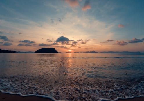 Golden and orange colors paint the sky as the sun sets over a serene beach. Gentle waves lap onto the shore, framed by distant islands and silhouettes of anchored boats.